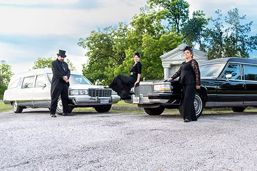Three people dressed in all black formal attire standing around the two hearses in a grave yard