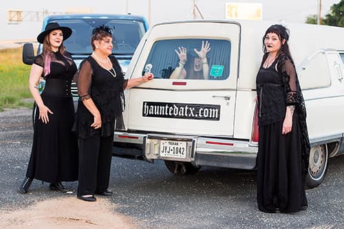 Three women standing outside of the white hearse while a fourth tries to claw her way out of the back of the hearse