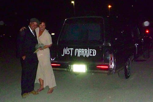A smiling couple standing behind the black hearse with a sign that says 'Just Married'