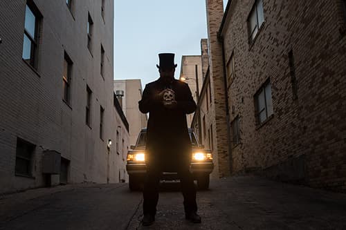 A man dressed in all black standing in an ally holding a skull with the hearse behind him